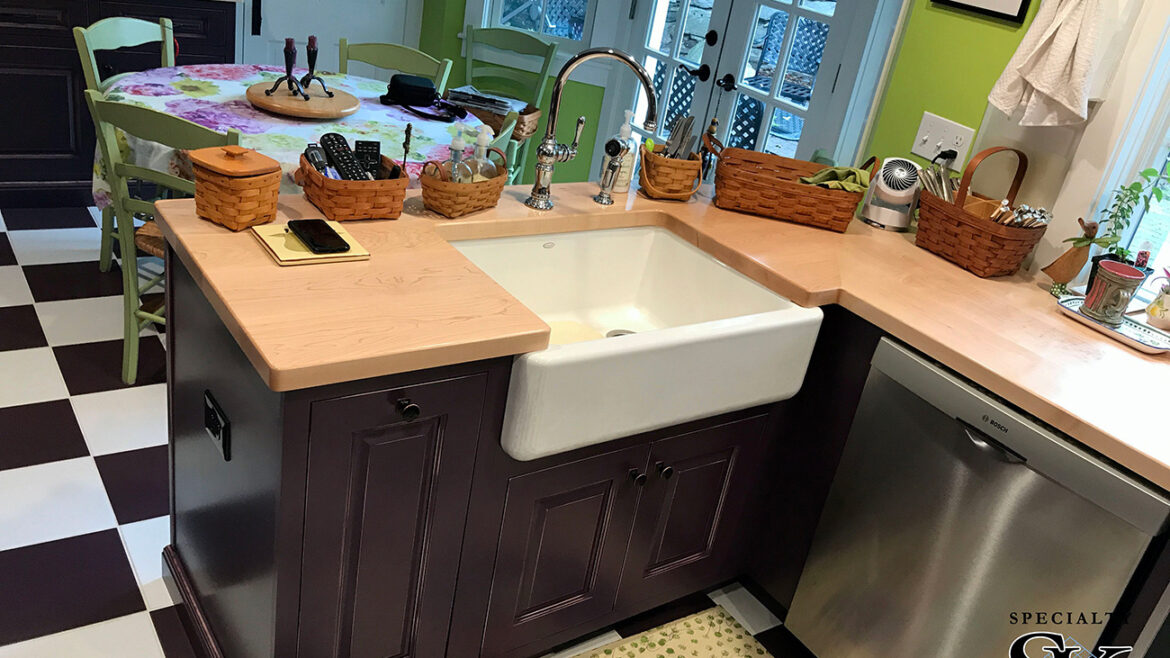 Cozy kitchen with green walls, checkered floor, farmhouse sink, wooden countertops, and table with floral tablecloth.