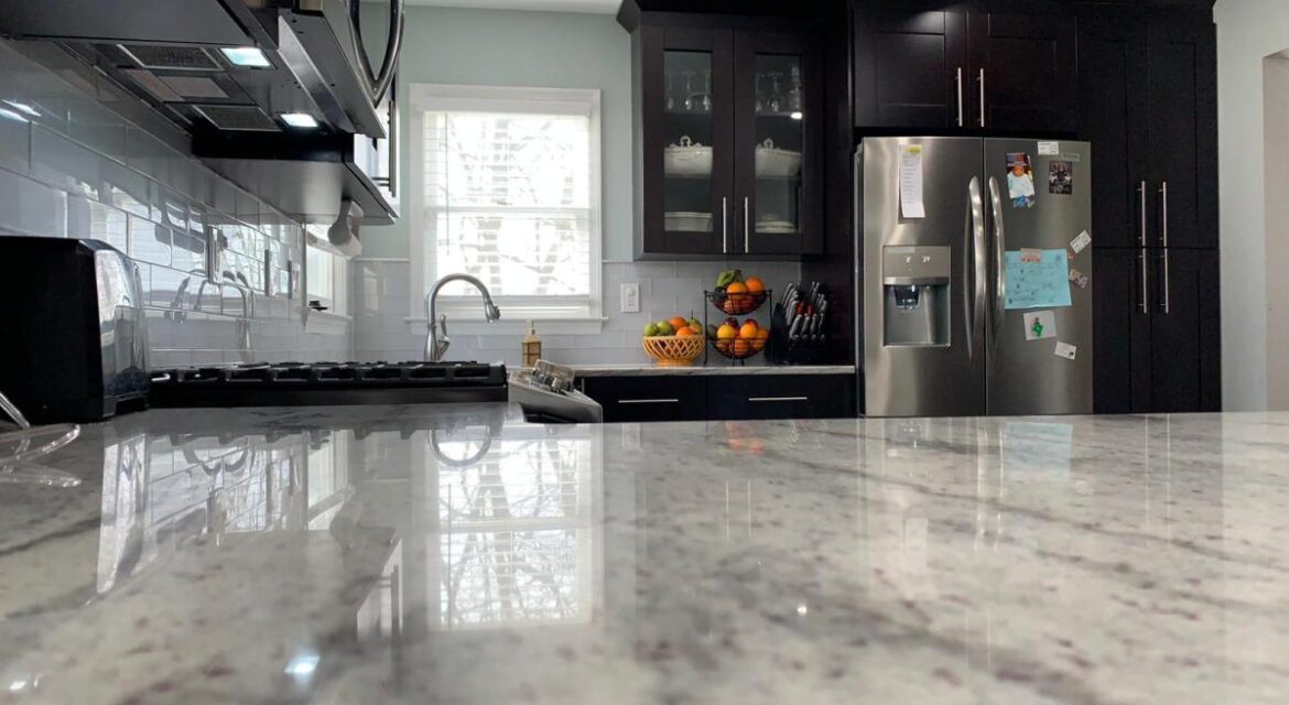 Modern kitchen with granite countertops, stainless steel appliances, dark cabinets, and a fruit bowl near the sink and fridge.