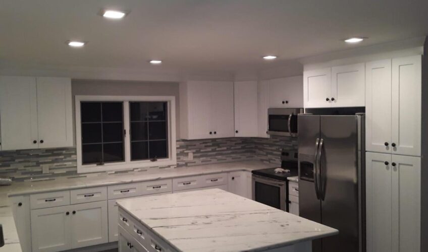 Modern kitchen with white cabinets, marble island, stainless steel appliances, and tiled backsplash under recessed lighting.