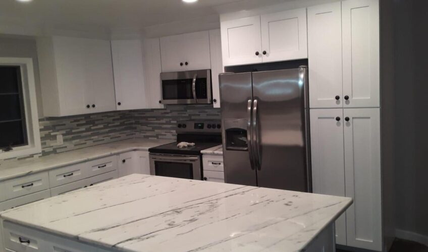 Modern kitchen with white cabinets, marble island, stainless steel appliances, and tiled backsplash under recessed lighting.