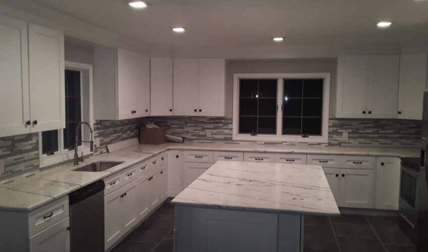 Modern kitchen with white cabinets, marble countertops, island, and tile backsplash under recessed lighting.