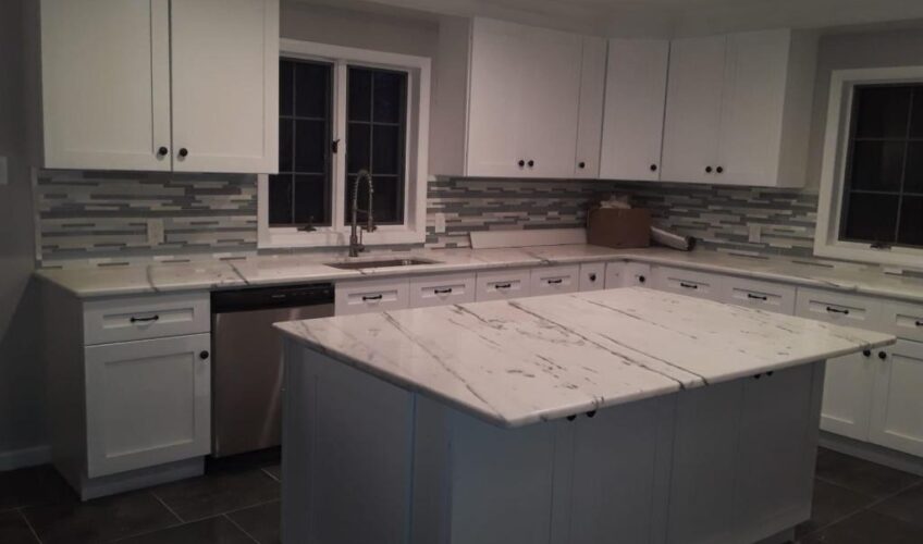 Modern kitchen with white cabinets, marble countertops, gray tile backsplash, and a large island under recessed lighting.