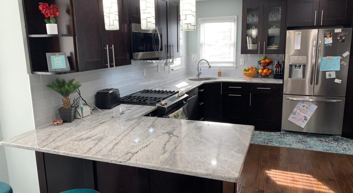 Modern kitchen with dark cabinets, marble countertops, stainless steel appliances, and teal barstools, brightly lit by windows.