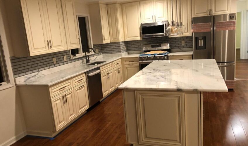 Modern kitchen with cream cabinets, marble countertops, stainless steel appliances, brick backsplash, and island on wooden floor.