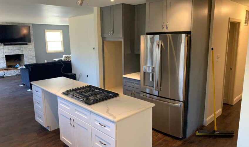 Modern kitchen with white island, built-in stovetop, gray cabinets, stainless steel fridge, and view into living area.