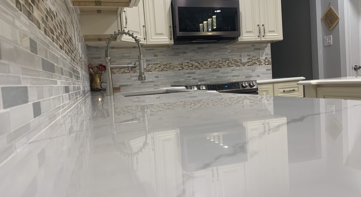 Modern kitchen with glossy white countertop, stainless steel faucet, mosaic tile backsplash, and white cabinets.