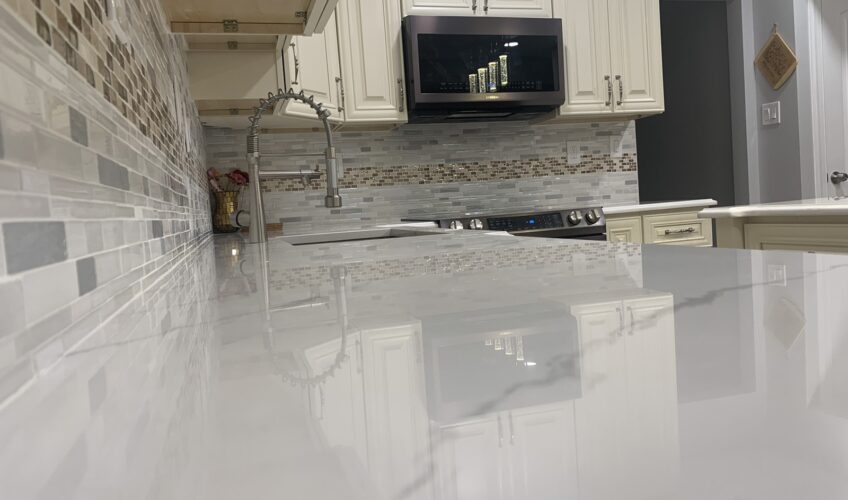 Modern kitchen with glossy white countertop, stainless steel faucet, mosaic tile backsplash, and white cabinets.