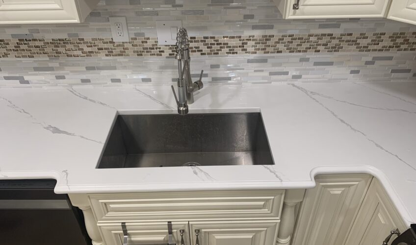 Modern kitchen sink with white marble countertop and mosaic tile backsplash, featuring cream cabinets and a chrome faucet.