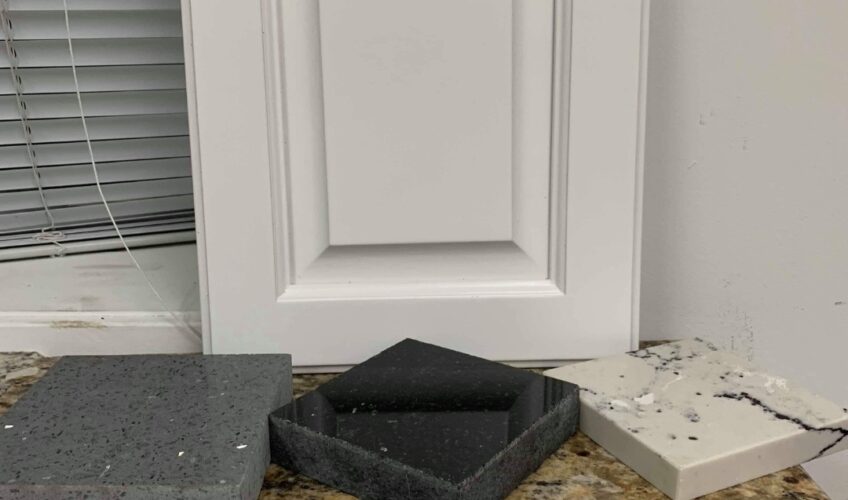 White cabinet door with gray and white stone samples on a granite countertop, next to a window with blinds.